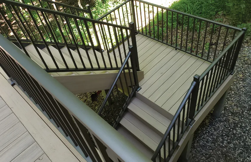 Tan composite deck stairs with landing and black aluminum railings surrounded by landscaping